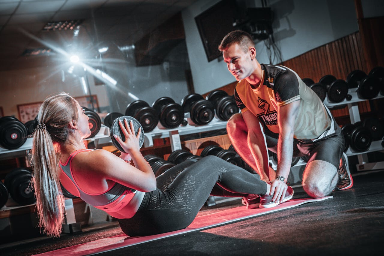 A personal trainer assists a woman doing sit-ups with a medicine ball in a gym setting.